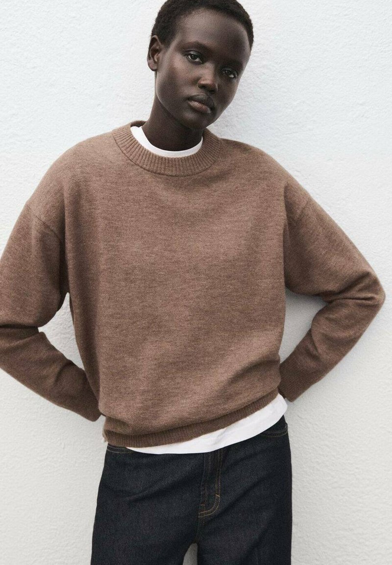 Maglione marrone lavorato a maglia con colletto rotondo, polsini e orlo a coste. Indossato sopra una t-shirt bianca, abbinato a jeans di denim scuro.