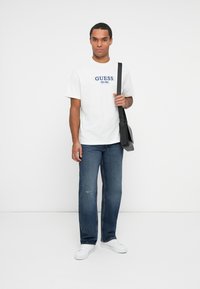 Camiseta de algodón blanca con logo azul de "GUESS", combinada con unos vaqueros de mezclilla azul oscuro. El modelo sostiene un bolso bandolera negro y viste zapatillas blancas.