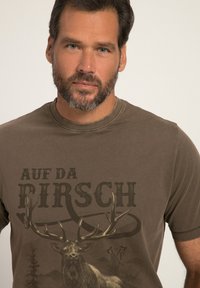 Camiseta marrón de manga corta con un gran gráfico de un ciervo con astas, texto que dice "AUF DA BIRSCH" y cuello acanalado.