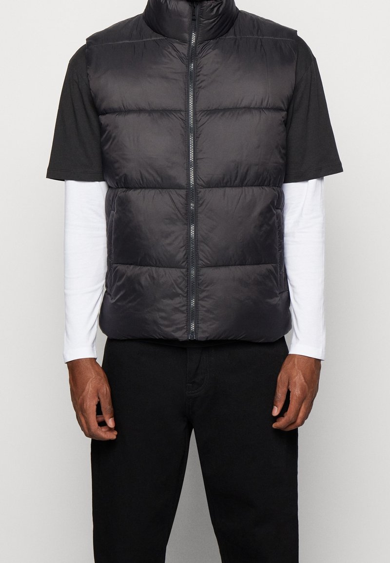 Osoba ubrana w czarną pikowaną kamizelkę bez rękawów zapinaną na zamek, nałożoną na czarną koszulkę z krótkim rękawem, pod którą znajduje się biała koszulka z długim rękawem.