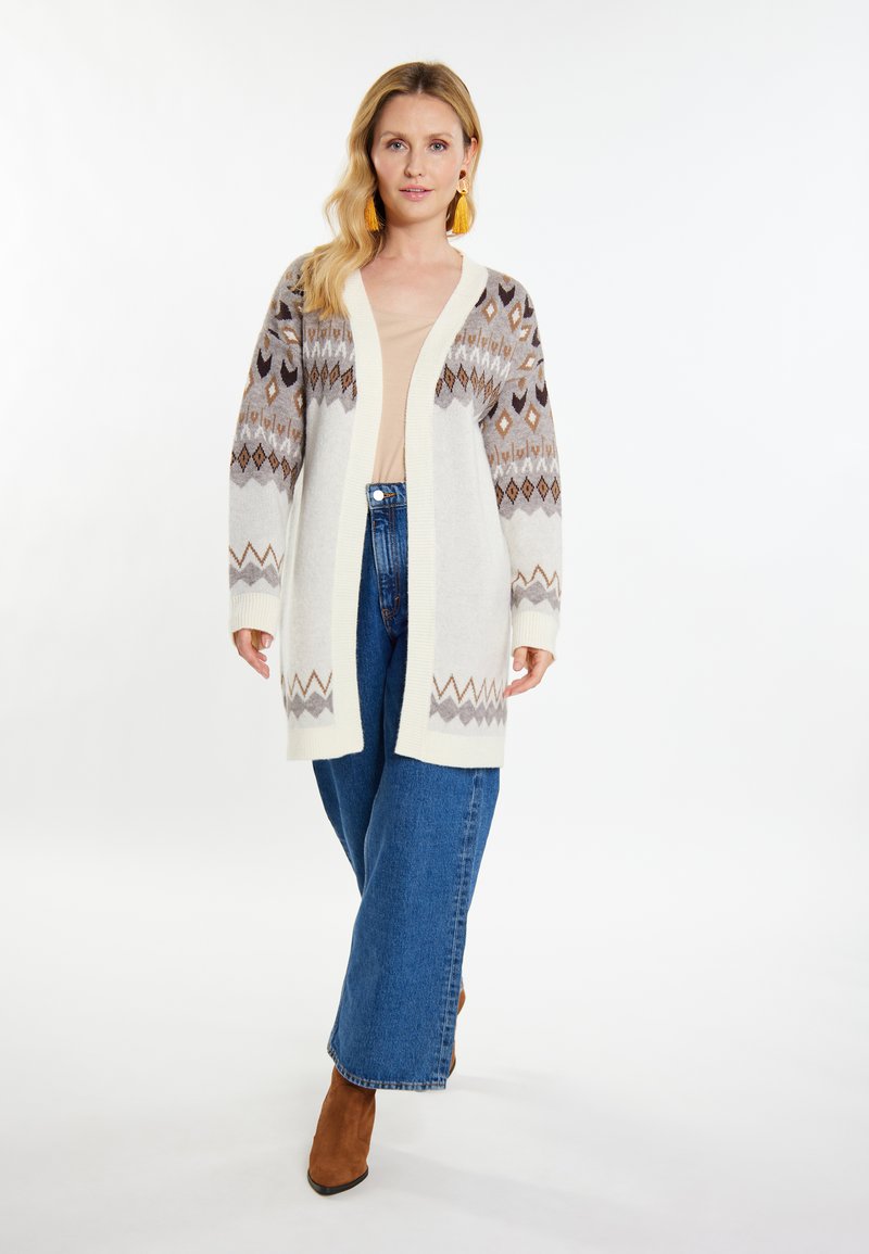 Langer, elfenbeinfarbener Cardigan mit braunen und grauen geometrischen Mustern, gerippten Bündchen und offener Vorderseite, kombiniert mit blauen weiten Jeans und braunen Stiefeln.