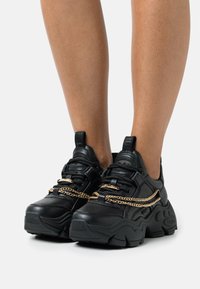 Black sneakers with a chunky sole, featuring a textured leather upper and metallic gold chain detail across the laces.