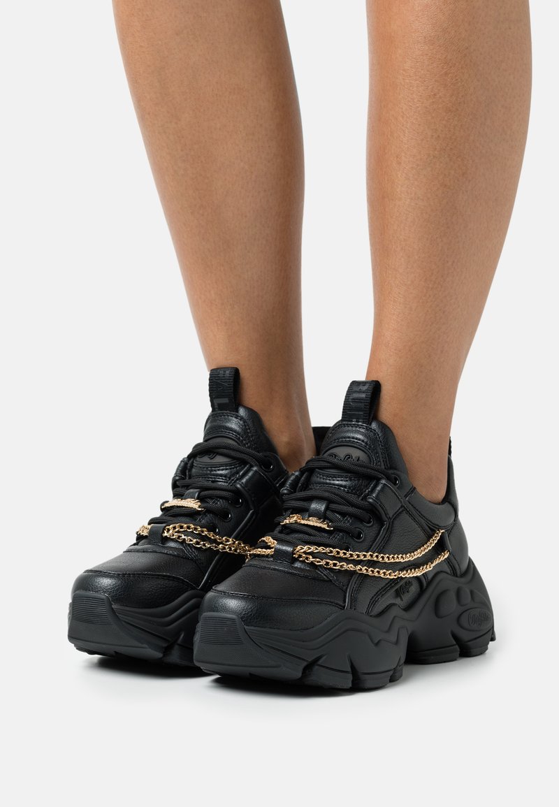 Black sneakers with a chunky sole, featuring a textured leather upper and metallic gold chain detail across the laces.