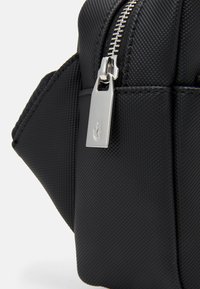 Borsa nera sintetica con una superficie testurizzata, caratterizzata da una zip in argento prominente e un'etichetta metallica con logo. Design compatto con bordi arrotondati.