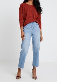 Pull rouge texturé surdimensionné à manches trois-quarts, associé à un jean taille haute bleu clair aux ourlets effilochés et des talons noirs à brides.