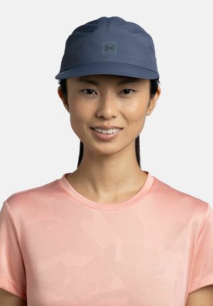 Joven mujer con gorra Buff azul oscuro y camiseta de manga corta texturizada rosa claro, sonriendo sobre un fondo liso.