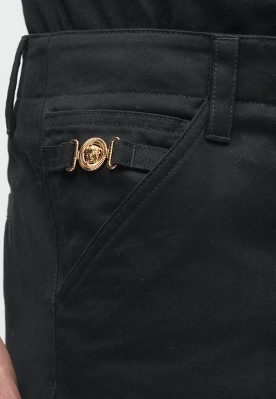 Fekete szövetnadrág jellegzetes arany oroszlán-emblémás gombbal, szögletes zsebekkel és sima textúrával.