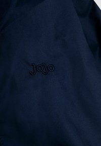Tumedat kangast, millel on sile tekstuur ja broderitud "Jojo" logo, mis on paigutatud silmapaistvalt veidi heledamas lõngavärvis.