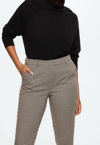 Maglione nero a collo alto abbinato a pantaloni a vita alta in plaid con motivo beige e marrone a pied de poule, dotati di tasche laterali e di un tessuto morbido.