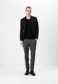 Zwarte blazer over een zwarte trui, grijze op maat gemaakte broek en zwarte enkellaarsjes. De outfit heeft een slim fit en subtiele texturen.