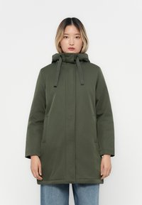 Manteau à capuche vert olive avec une coupe droite, cordons de réglage et deux poches latérales, associé à un jean bleu à jambes larges.