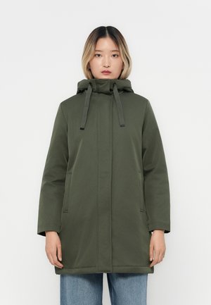 Manteau à capuche vert olive avec une coupe droite, cordons de réglage et deux poches latérales, associé à un jean bleu à jambes larges.