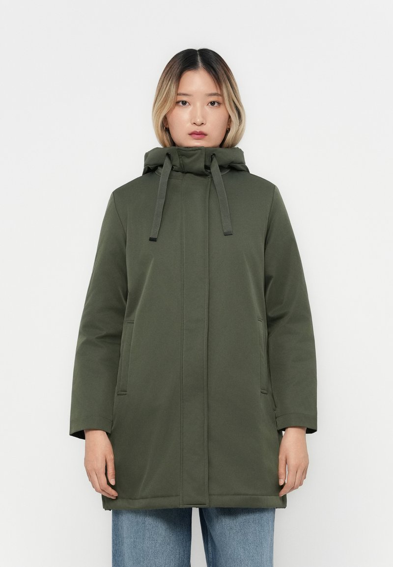 Manteau à capuche vert olive avec une coupe droite, cordons de réglage et deux poches latérales, associé à un jean bleu à jambes larges.