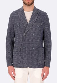 Blazer doppiopetto blu navy realizzato in tessuto testurizzato, con tasche laterali e dettagli in bottone. Indossato sopra una camicia chiara.