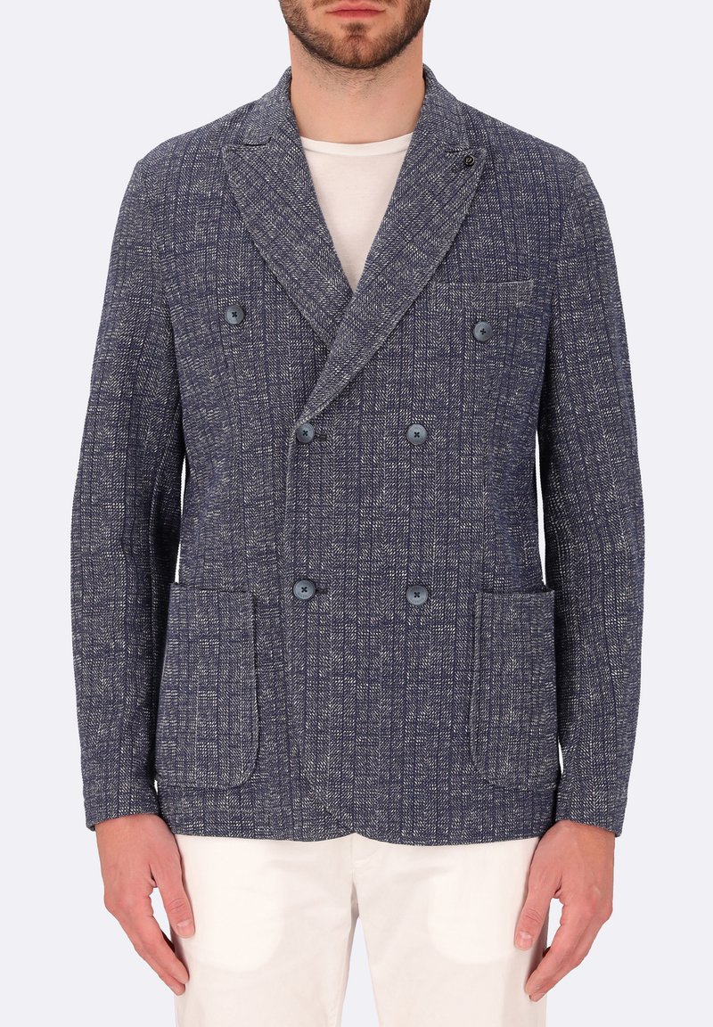 Blazer doppiopetto blu navy realizzato in tessuto testurizzato, con tasche laterali e dettagli in bottone. Indossato sopra una camicia chiara.