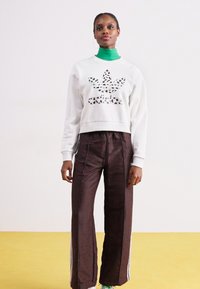 Hvit sweatshirt med utskåret leo-punkts logo, kombinert med mørkebrune satengbukser med hvite striper langs sidene. Grønn undertrøye synlig.