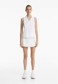 Débardeur blanc sans manches avec un col en V, en tissu texturé, associé à une jupe blanche avec des fentes latérales et des bordures contrastantes.
