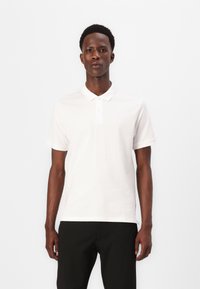 Polo bianco con colletto classico, maniche corte e patta con due bottoni. Abbinato a pantaloni neri a righe sottili.