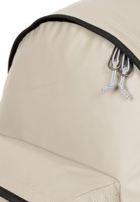 Zaino beige chiaro con un esterno testurizzato e resistente, dotato di una chiusura superiore a cerniera e due tiranti metallici con dettagli in corda grigia.