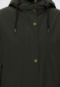 Veste à capuche vert foncé avec cordons marron et boutons-pression en laiton sur le devant, gros plan sur le haut du torse et la zone du cou.