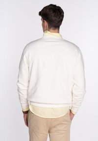Sudadera color crema con una textura suave, cuello redondeado y puños acanalados, superpuesta sobre una camisa de color amarillo claro con cuello.