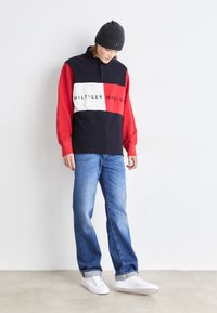 Rot und marineblau farblich abgeblockter Sweatshirt mit Logo-Text, kombiniert mit hellblauen Jeans und weißen Sneakern. Eine graue Strickmütze rundet den Look ab.