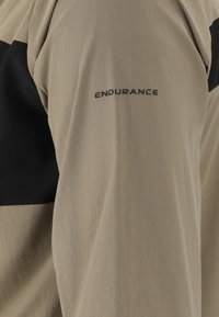 Helles beiges Langarmshirt mit einem schwarzen Panel. Verfügt über einen strukturierten Stoff und ein "ENDURANCE"-Logo am Ärmel.