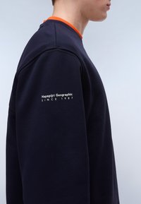 Granatowy sweter z pomarańczowym ściągaczem pod szyją. Zawiera biały napis "Napapijri Geographic Since 1987" na lewym rękawie. Miękka tekstura.