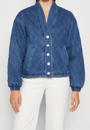 Vrouw draagt een blauwe gewatteerde jas met knoopsluiting en elastische boorden, gecombineerd met een witte broek tegen een effen achtergrond.