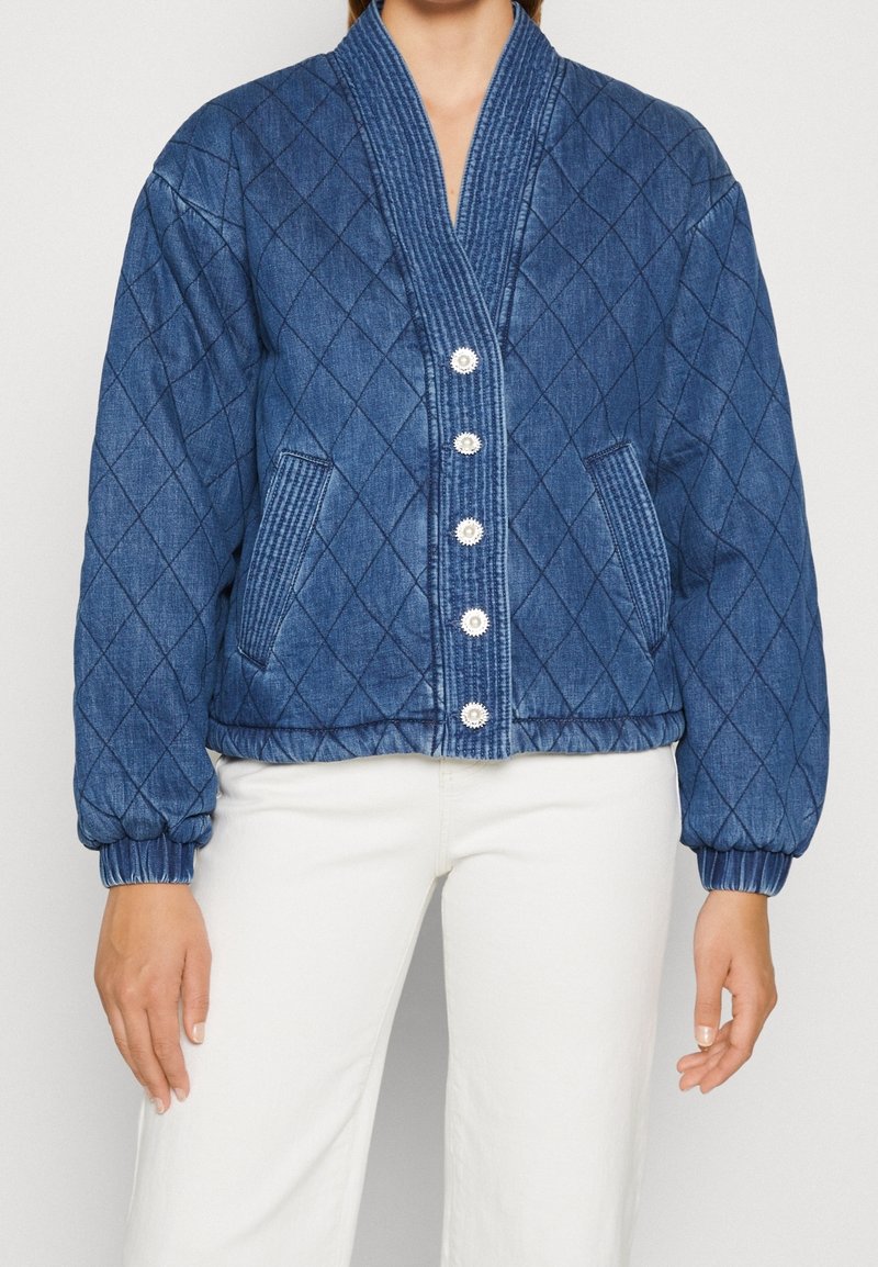 Femme portant une veste matelassée bleue avec fermeture à boutons et poignets élastiques, associée à un pantalon blanc sur un fond uni.