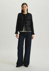 Veste noire texturée avec des boutons argentés, deux poches avant et des détails en franges, portée sur un haut rayé et assortie avec des jeans évasés.