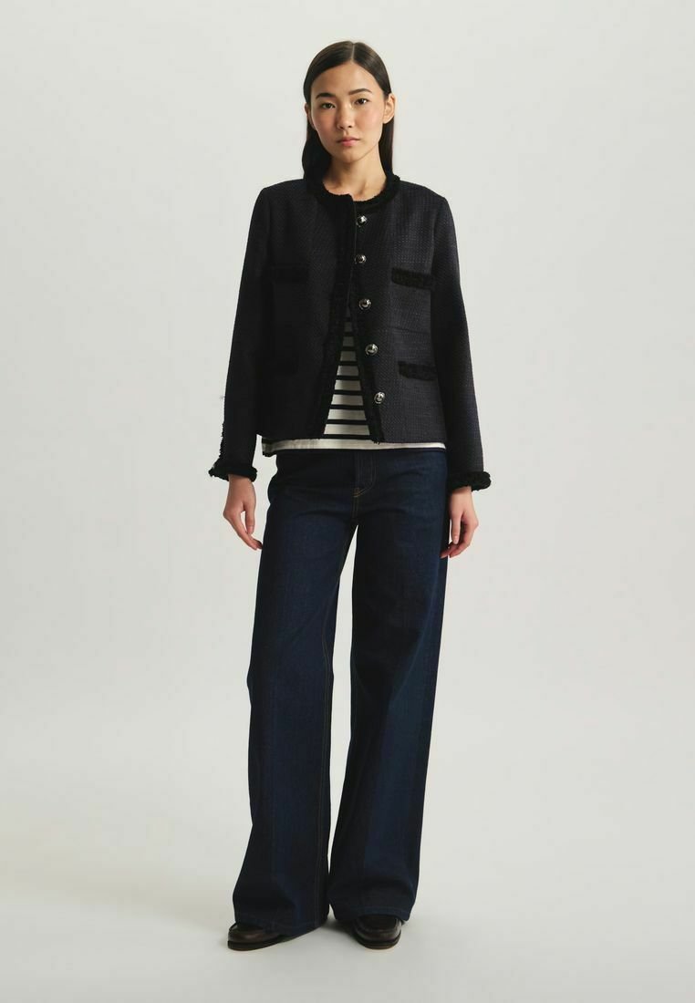 Veste noire texturée avec des boutons argentés, deux poches avant et des détails en franges, portée sur un haut rayé et assortie avec des jeans évasés.