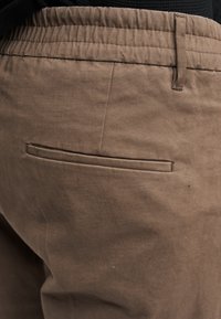 Pantalon en coton marron avec une ceinture élastique visible et une poche arrière discrète. Le tissu présente une finition lisse et légèrement texturée.