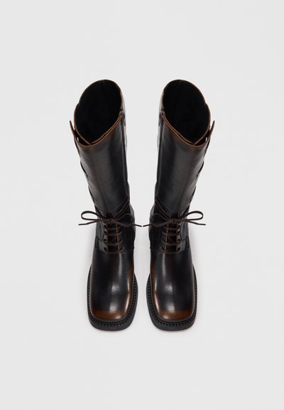 Brown leather knee-high boots with a glossy finish, lace-up front, and contrasting black texture panel, featuring a stitched sole and open top.