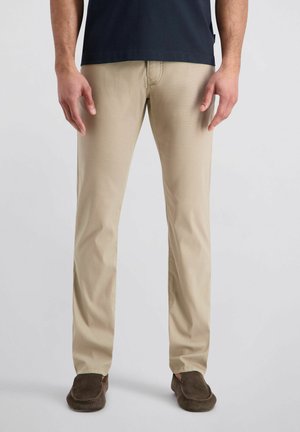 Homme portant un pantalon beige à jambes droites, une chemise bleu marine foncé et des chaussures marron sans lacets, debout devant un fond gris clair.