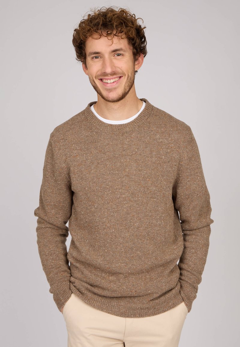 Pull en tricot marron avec des éclats clairs, col rond et poignets côtelés. Porté sur une chemise blanche, associé à un pantalon de couleur claire.