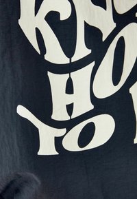 Textil med stora, djärva, vita bokstäver på en mörk bakgrund. Tyget verkar mjukt med en slät yta. Texten inkluderar ord som är skurna i lekfulla former.