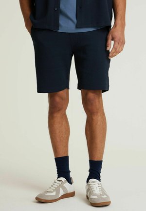 Donkerblauwe gemarmerde shorts met een aansluitende pasvorm, gecombineerd met een lichtblauw shirt, donkerblauwe geribbelde sokken en grijze sneakers met witte accenten.