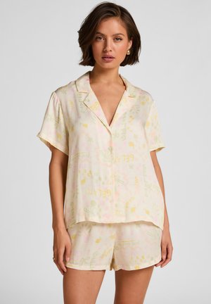 Vrouw die een licht pastelkleurig shirt met korte mouwen en knopen draagt, gecombineerd met bijpassende korte broek met subtiele bloemen- en tekstpatronen, staand tegen een effen achtergrond.