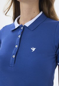 Koninklijk blauwe polo met een witte kraag, voorzien van een knoopsluiting aan de voorkant en een geborduurd vogel-logo aan de linkerkant. Gewatteerde stof.