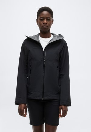 Oseba, ki nosi črno jakno z zadrgo in sivo kapuco nad belo majico in črnimi kratkimi hlačami, stoji pred preprostim ozadjem.