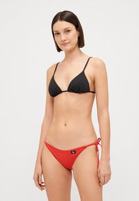 Haut de bikini en triangle noir avec fines bretelles, associé à un bas de bikini rouge à nouer sur les côtés arborant une petite étiquette logo. Tissu lisse.