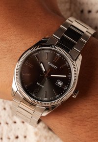 Montre en acier inoxydable argenté avec un cadran gris foncé, des marqueurs d'heure lumineux, un affichage de la date et une lunette polie. Résistante à l'eau jusqu'à 30 mètres.