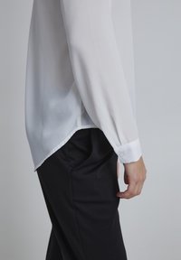 White, sheer blouse with long sleeves, slight sheen, and a curved hem. Worn over black, fitted trousers with pocket detail.