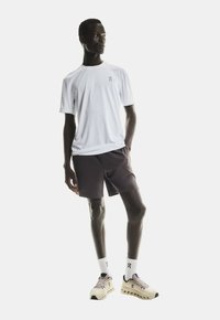 Männlicher Model trägt ein weißes Kurzarmhemd, dunkle Shorts, weiße Marken-Socken und beige Sneaker und steht mit einer Hand in der Tasche.
