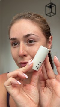 ILIA huidverzorgingsproduct in een cilindervormige witte container met matte afwerking en een zwart logo. Model houdt het dicht bij haar gezicht, waarbij haar nagels zichtbaar zijn.