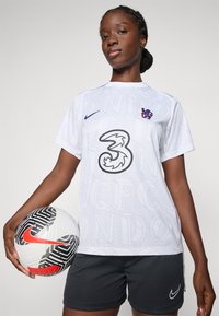 Witte short-sleeve voetbalshirt met '3' logo, subtiel patroon en blauwe clubcrest. Zwarte korte broek en een zwart-wit geblokte voetbal.