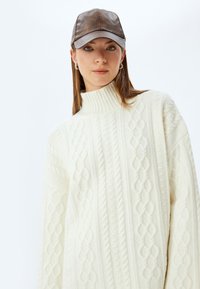 Crème-kleurige, kabelgebreide turtleneck trui met textuurpatronen, gecombineerd met een bruine leren baseballpet met grijze accenten.