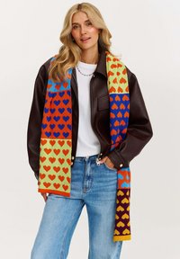 Farebný pletený šál s motívom sŕdc v červenej, modrej, oranžovej a žltej farbe, vyrobený z mäkkej látky, prehodený cez čiernu koženú bundu.