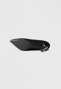 Zapato de tacón negro de punta afilada con un exterior texturizado, un tacón negro brillante y una silueta esbelta. Suela lisa con un ligero diseño en estrías.